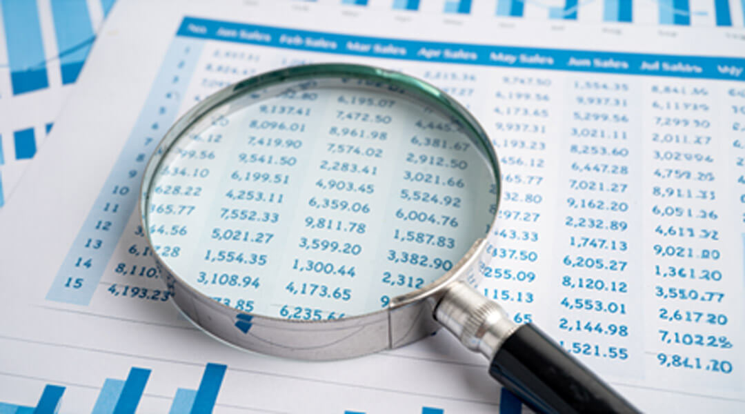 A magnifying glass rests on financial spreadsheets, enlarging a section of numbers for closer examination.